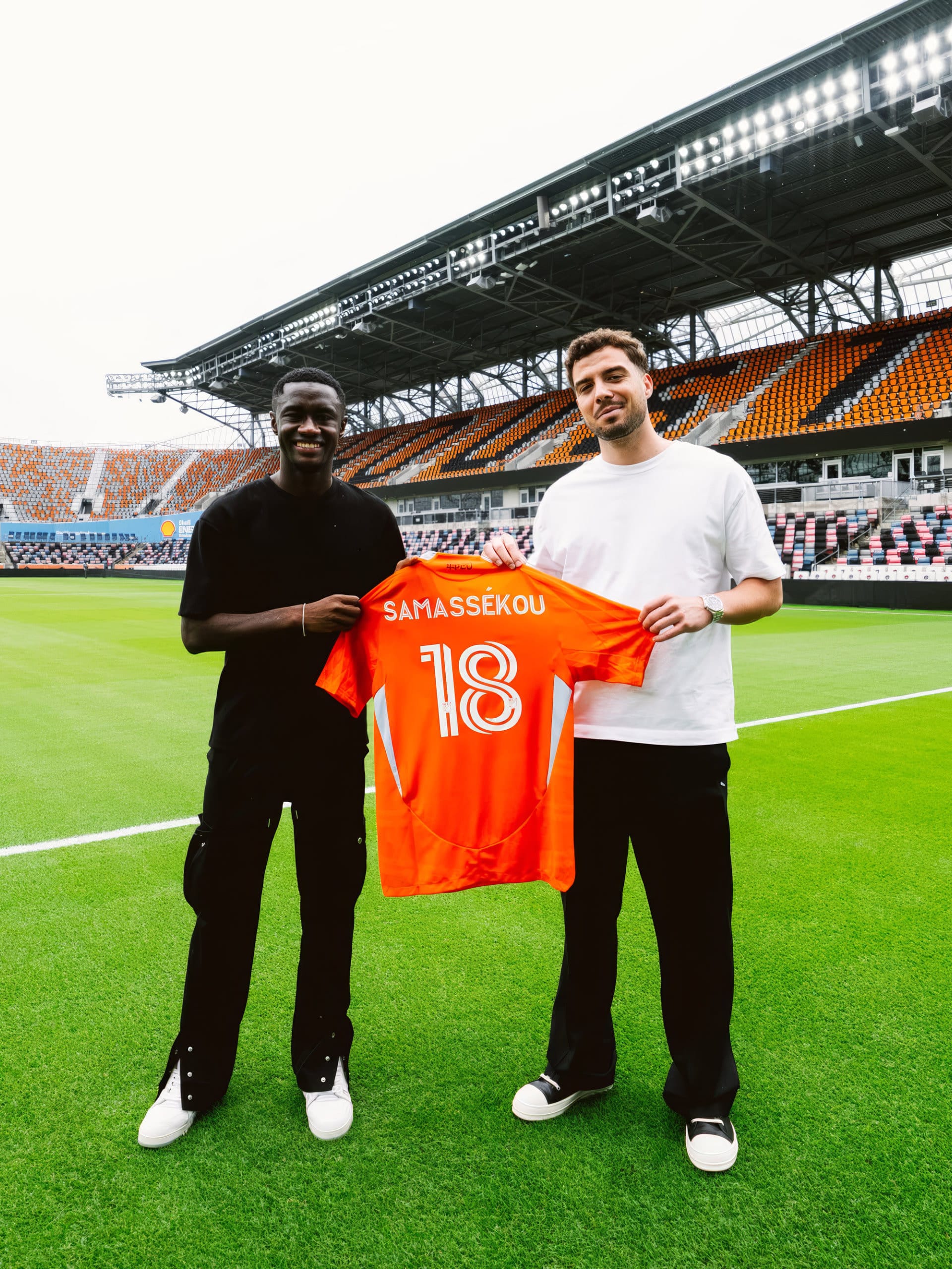 Diadié Samassékou und Felix Lang auf dem Rasen eines leeren Stadions, gemeinsam ein orangefarbenes Trikot mit der Nummer 18 haltend